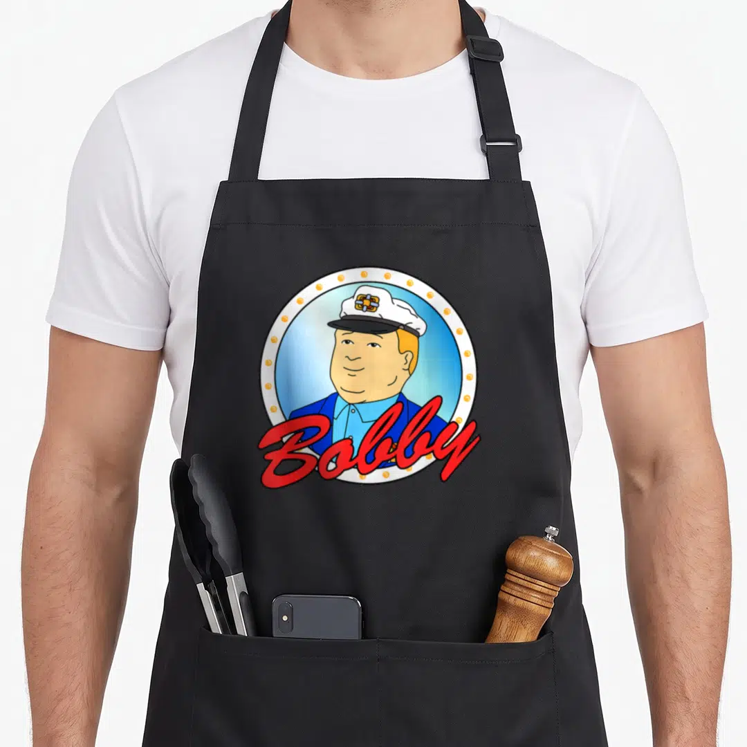 Sailor cartoon apron - Captain portrait in white circle with orange dots and red Bobby script — shown on a black cotton canvas apron against a white background