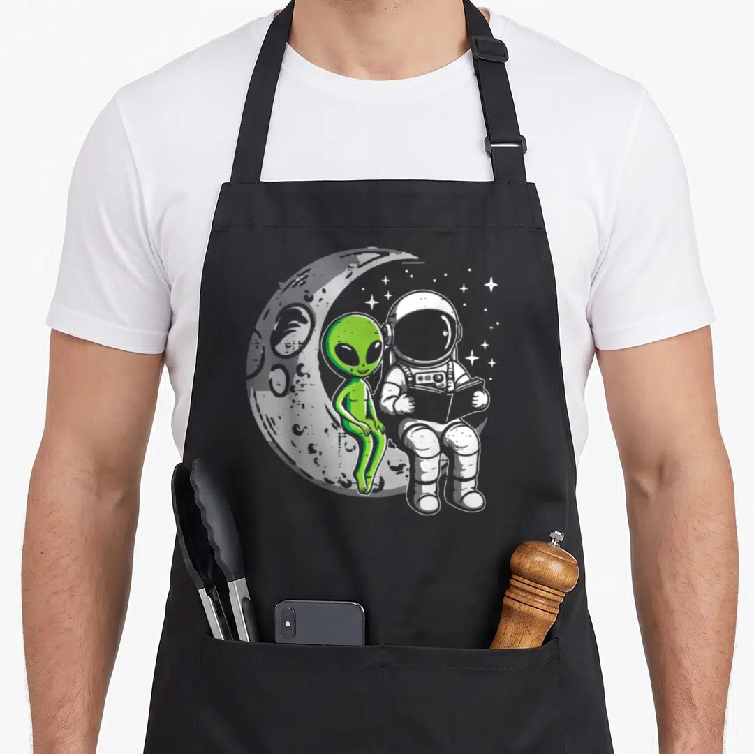 Astronaut mom space apron - Green alien and grey astronaut sitting on a crescent moon reading a book with white stars — shown on a black cotton canvas apron against a white background