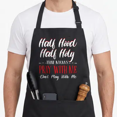 Holy novelty apron - Stacked white and red text reading Half Hood Half Holy Pray With Me with arrow and heart graphics — shown on a black cotton canvas apron against a white background