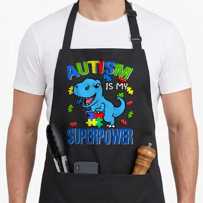 Superpower autism dad apron - Blue cartoon dinosaur holding multicolored puzzle piece with arched text reading AUTISM — shown on a black cotton canvas apron against a white background