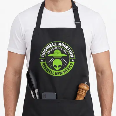 Roswell aviation apron - Lime green alien head and flying saucer inside circular emblem with established 1947 text — shown on a black cotton canvas apron against a white background