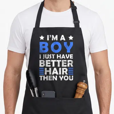Cute better hair apron - Stacked white and blue text reading I'M A BOY I JUST HAVE BETTER HAIR THEN YOU flanked — shown on a black cotton canvas apron against a white background