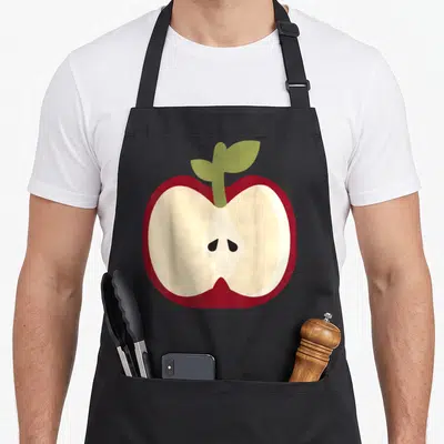 Easy apple apron - Cartoon sliced red apple with cream interior and green stem on black background — shown on a black cotton canvas apron against a white background