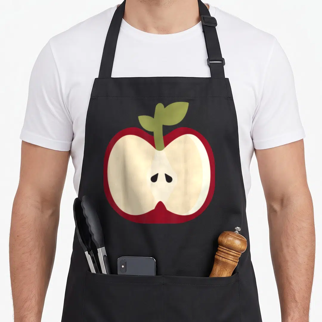 Easy apple apron - Cartoon sliced red apple with cream interior and green stem on black background — shown on a black cotton canvas apron against a white background