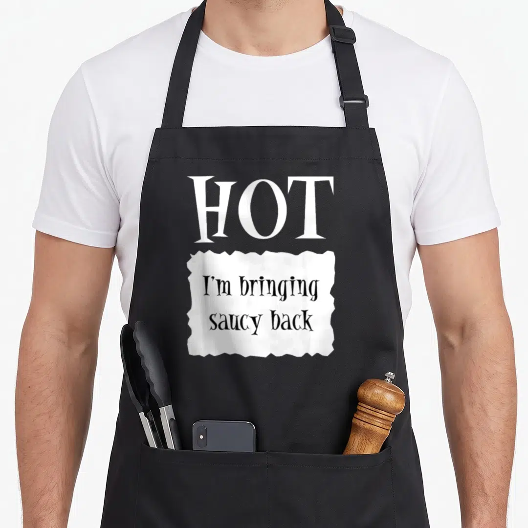 Hot packet halloween apron - White HOT text above torn paper note reading I'm bringing saucy back in black font — shown on a black cotton canvas apron against a white background