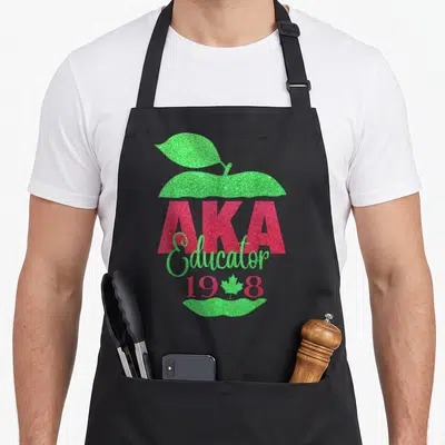 Pretty teacher apron - Red AKA Educator 1908 text over green apple with maple leaf — shown on a black cotton canvas apron against a white background