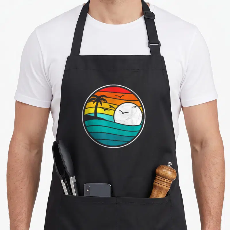 Beach 80s apron - Circular retro sunset with striped red orange yellow teal sky, white sun, palm tree, and waves — shown on a black cotton canvas apron against a white background