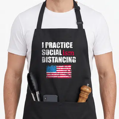 Anti political aprons - Stacked white and red text reading I Practice Socialism Distancing above a distressed USA flag — shown on a black cotton canvas apron against a white background