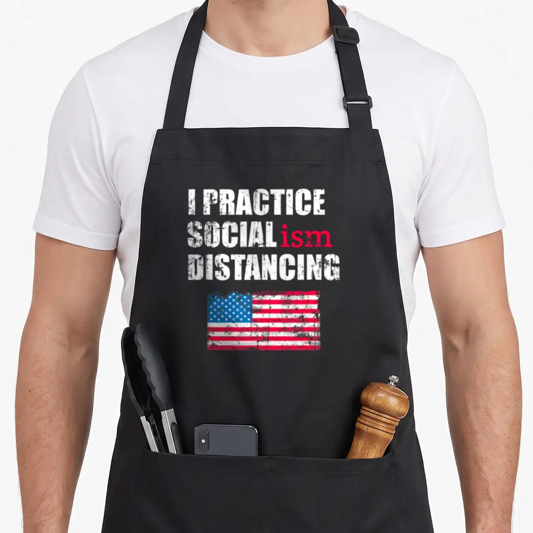 Anti political aprons - Stacked white and red text reading I Practice Socialism Distancing above a distressed USA flag — shown on a black cotton canvas apron against a white background