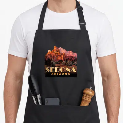 Arizona sedona apron - Realistic red rock mountain landscape with stacked SEDONA ARIZONA text in orange and yellow — shown on a black cotton canvas apron against a white background