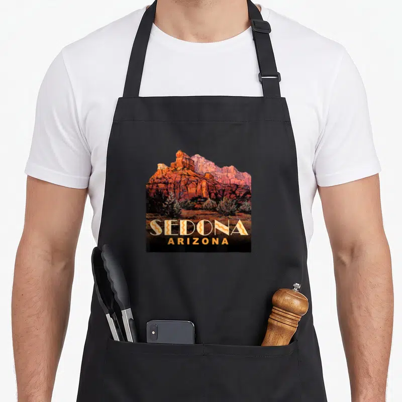 Arizona sedona apron - Realistic red rock mountain landscape with stacked SEDONA ARIZONA text in orange and yellow — shown on a black cotton canvas apron against a white background