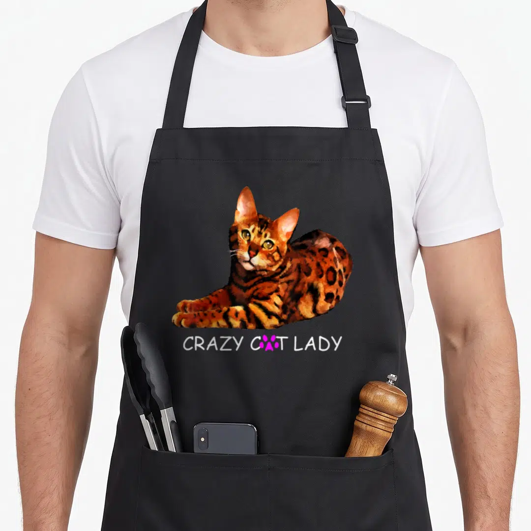 Colorful crazy cat lady apron - Realistic orange spotted cat above white text with pink paw print replacing the letter — shown on a black cotton canvas apron against a white background