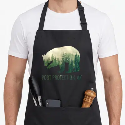 Port bear apron - Bear silhouette filled with green pine trees and fog above PORT PROTECTION, AK text — shown on a black cotton canvas apron against a white background