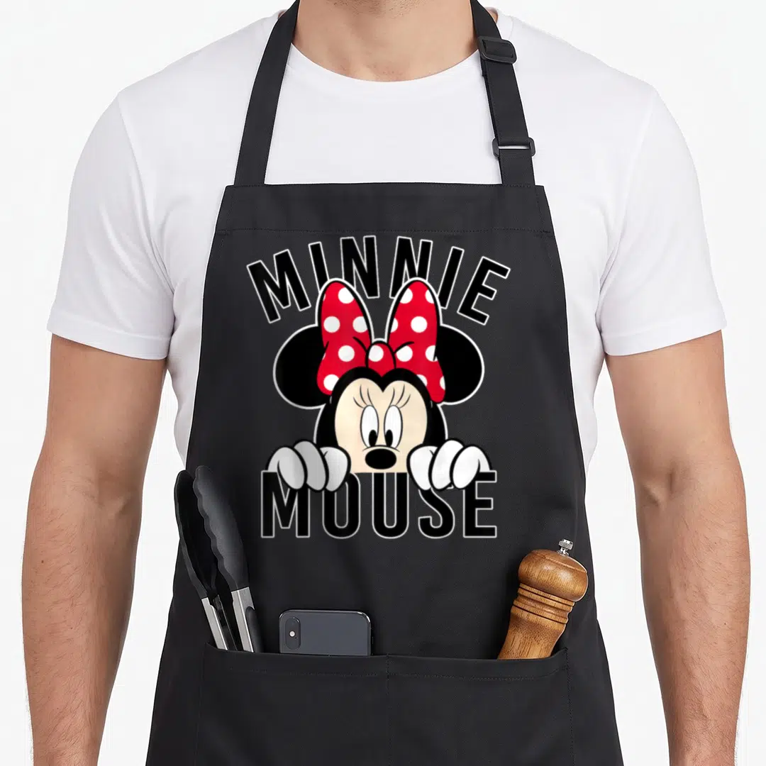 Rock dot apron - Minnie Mouse character peeking from behind black MINNIE MOUSE text with red polka dot bow — shown on a black cotton canvas apron against a white background