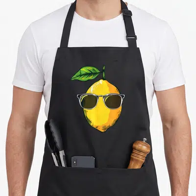 Sunglass lemon apron - Cartoon yellow lemon wearing white rimmed sunglasses with a green leaf attached to the top — shown on a black cotton canvas apron against a white background