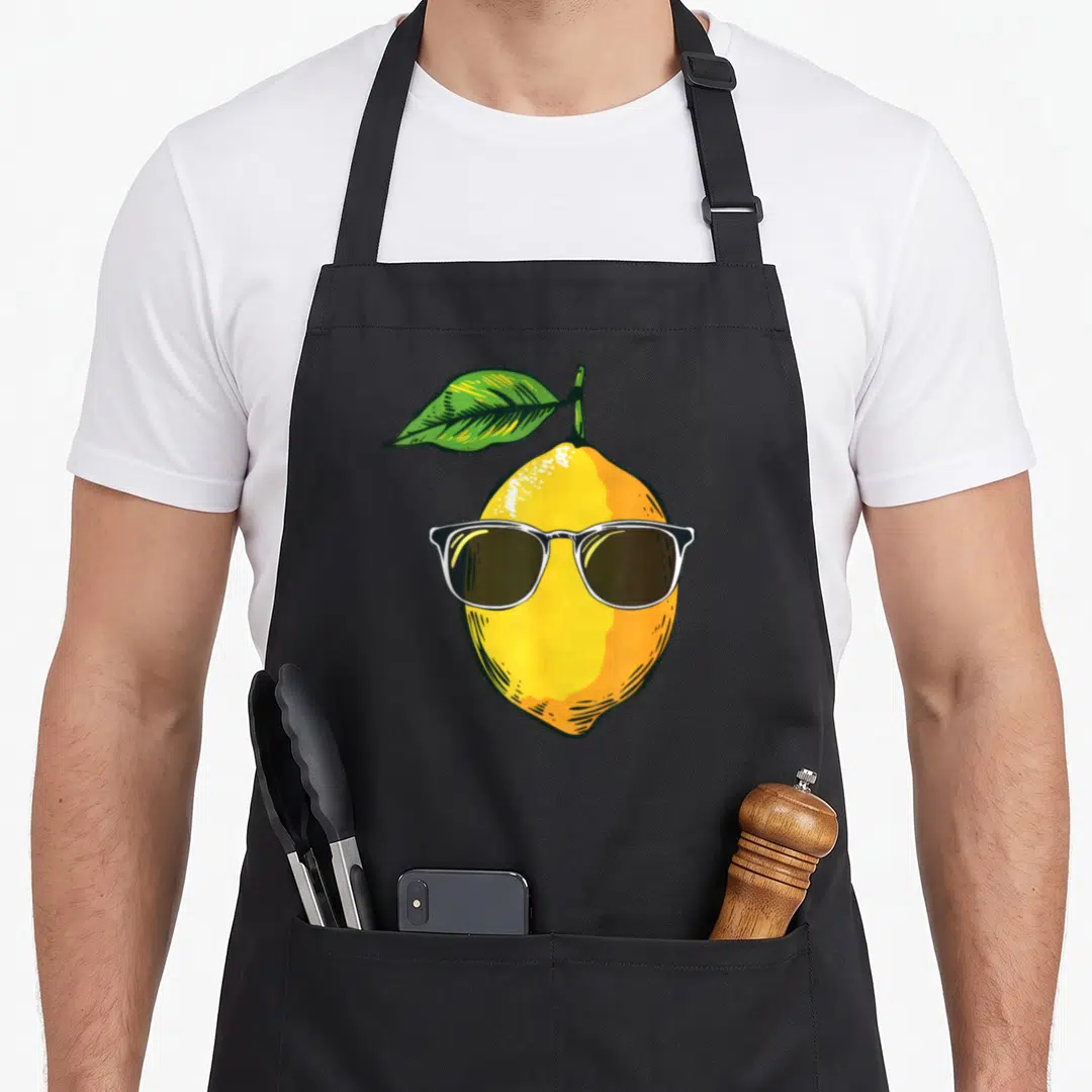 Sunglass lemon apron - Cartoon yellow lemon wearing white rimmed sunglasses with a green leaf attached to the top — shown on a black cotton canvas apron against a white background