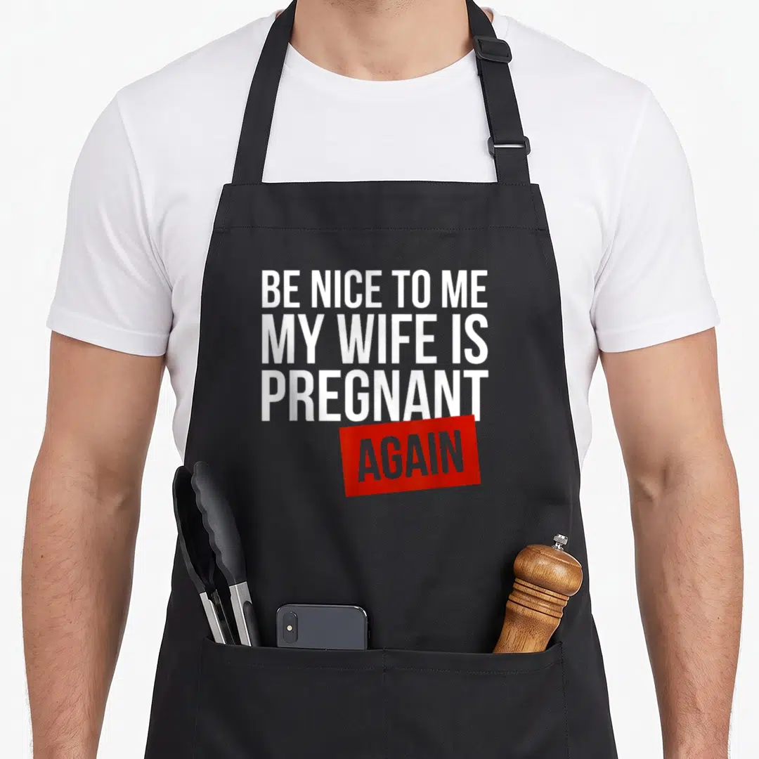 Funny pregnant wife apron - Stacked white text reading BE NICE TO ME MY WIFE IS PREGNANT AGAIN inside a red tilted — shown on a black cotton canvas apron against a white background