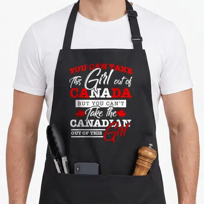 Maple leaf canadian aprons - Stacked red and white text You Can Take This Girl Out of Canada with two maple leaves — shown on a black cotton canvas apron against a white background