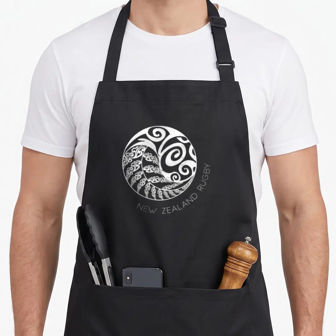 Ying new zealand apron - White circular Maori swirl graphic with curved text NEW ZEALAND RUGBY below — shown on a black cotton canvas apron against a white background
