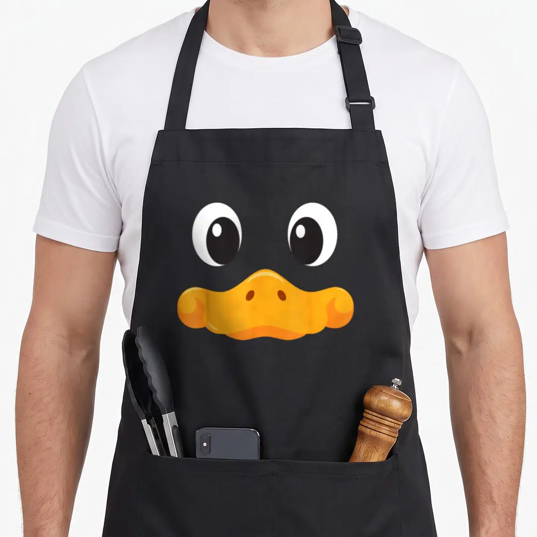 Duck funny halloween apron - Cartoon duck face with two large white eyes and wide orange beak — shown on a black cotton canvas apron against a white background