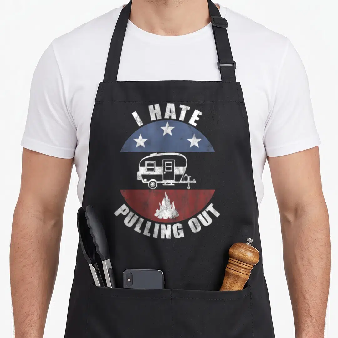Pulling travel trailer apron - Distressed white text I HATE PULLING OUT over blue stars and red fire with a camper — shown on a black cotton canvas apron against a white background