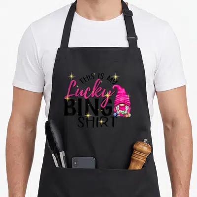 Gnome bingo apron - Pink gnome holding five bingo balls next to stacked text reading This Is My Lucky Bingo Shirt — shown on a black cotton canvas apron against a white background