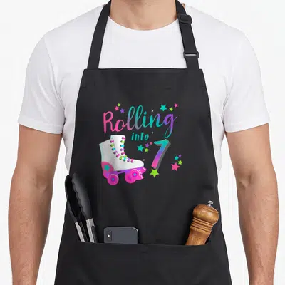 Decade birthday apron - Rolling into 7 text with white roller skate and scattered stars in pink blue and green — shown on a black cotton canvas apron against a white background