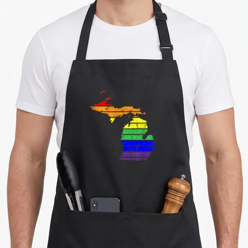 Distressed michigan gay pride apron - Rainbow striped Michigan state map silhouette on black fabric background — shown on a black cotton canvas apron against a white background