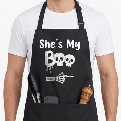 Halloween couple apron - White distressed text She's My Boo with two skulls and pointing skeleton hand — shown on a black cotton canvas apron against a white background