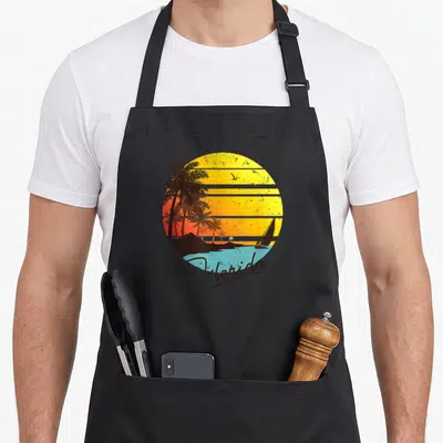 State summer aprons - Retro Florida sunset with striped sun, palm trees, sailboat, and script text — shown on a black cotton canvas apron against a white background