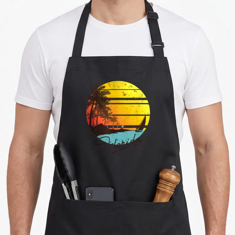 State summer aprons - Retro Florida sunset with striped sun, palm trees, sailboat, and script text — shown on a black cotton canvas apron against a white background