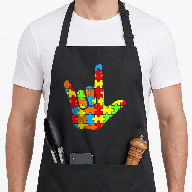 Autism asl sign for apron - Cartoon hand filled with red yellow blue green orange interlocking puzzle pieces — shown on a black cotton canvas apron against a white background