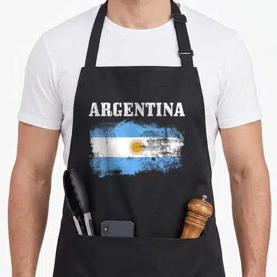 Argentina flag pride apron - Distressed Argentina flag print with light blue white yellow stripes and sun emblem — shown on a black cotton canvas apron against a white background