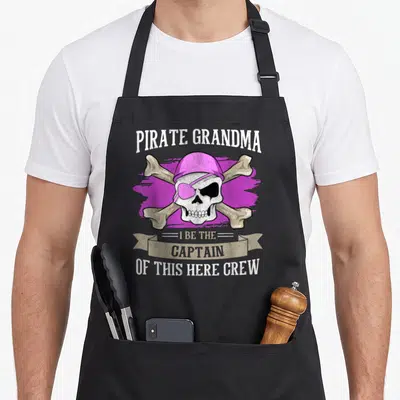 Pirate grandma apron - Pink hat skull with crossed bones and PIRATE GRANDMA text on purple brushstroke — shown on a black cotton canvas apron against a white background