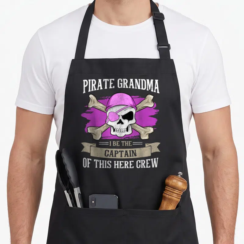 Pirate grandma apron - Pink hat skull with crossed bones and PIRATE GRANDMA text on purple brushstroke — shown on a black cotton canvas apron against a white background