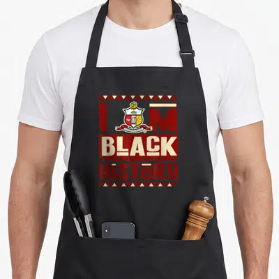Fraternity alpha apron - Red and cream text I M BLACK HiSTORY with a central shield crest and triangle borders — shown on a black cotton canvas apron against a white background