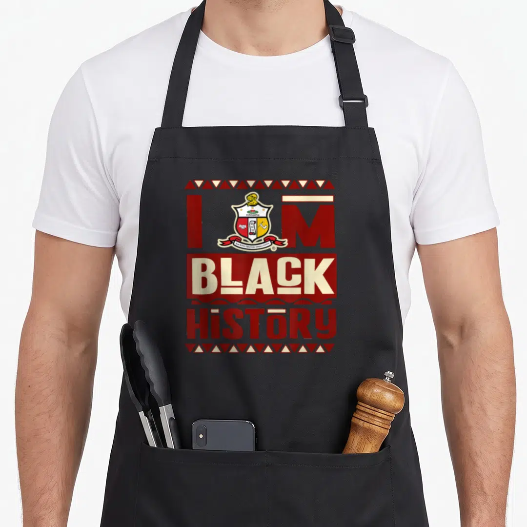 Fraternity alpha apron - Red and cream text I M BLACK HiSTORY with a central shield crest and triangle borders — shown on a black cotton canvas apron against a white background