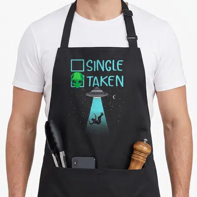 Single funny alien apron - Green alien head checking Taken box above UFO beaming up person silhouette against starry — shown on a black cotton canvas apron against a white background