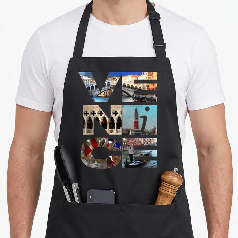 Trip italy apron - VENICE text formed by six photo collages of bridges, masks, and gondolas — shown on a black cotton canvas apron against a white background