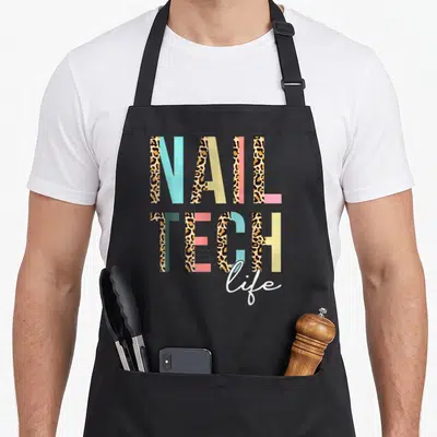 Leopard manicurist apron - NAIL TECH life text design with tan leopard print and colored blocks — shown on a black cotton canvas apron against a white background