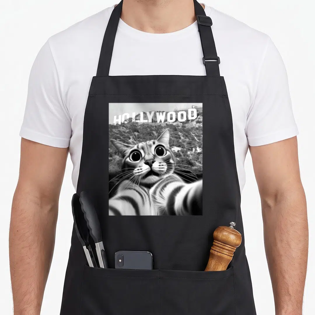 California cat apron - Black and white cat selfie photo with Hollywood sign text on hillside — shown on a black cotton canvas apron against a white background