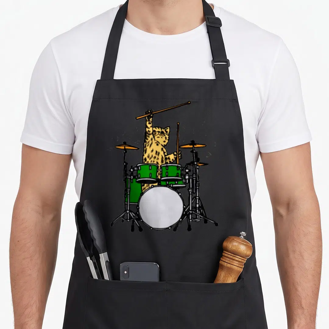 Drummer cute cat apron - Spotted yellow cartoon cat playing green drums with orange cymbals and floating notes — shown on a black cotton canvas apron against a white background