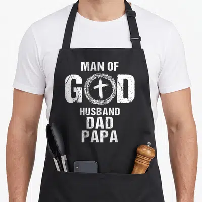 Husband papa father day apron - White distressed text MAN OF GOD HUSBAND DAD PAPA with cross inside crown of thorns — shown on a black cotton canvas apron against a white background