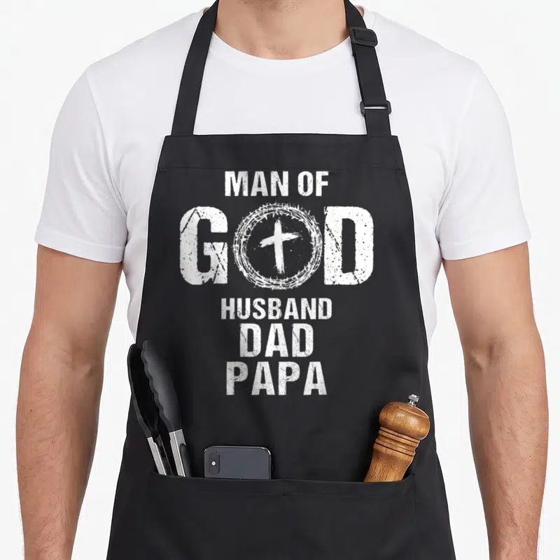 Husband papa father day apron - White distressed text MAN OF GOD HUSBAND DAD PAPA with cross inside crown of thorns — shown on a black cotton canvas apron against a white background