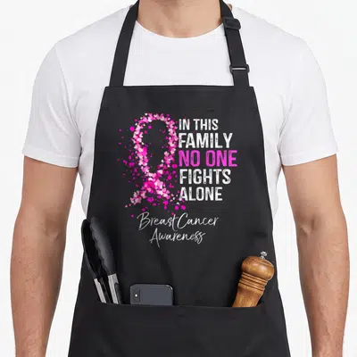 Family breast cancer awareness aprons - Pink ribbon formed by small hearts next to white and pink text reading IN THIS — shown on a black cotton canvas apron against a white background