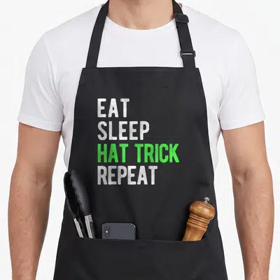 Eat snapback apron - Stacked text EAT SLEEP HAT TRICK REPEAT in white and green block letters — shown on a black cotton canvas apron against a white background