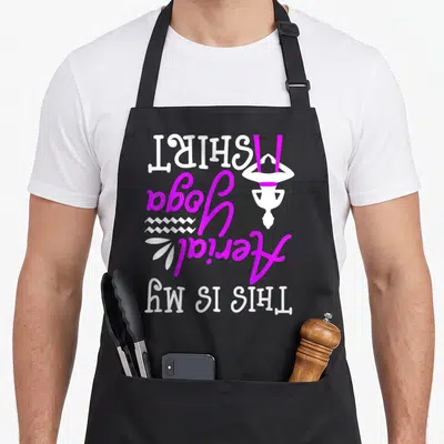Aerial grill instructor apron - Vertical white and purple THIS IS MY AERIAL YOGA SHIRT text with inverted figure — shown on a black cotton canvas apron against a white background