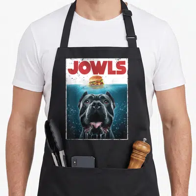 Jowl cane apron - Red JOWLS text above black dog underwater reaching for floating burger — shown on a black cotton canvas apron against a white background
