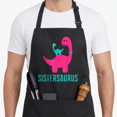 Sistersaurus themed aprons - Pink and cyan cartoon dinosaurs with SISTERSAURUS text below — shown on a black cotton canvas apron against a white background