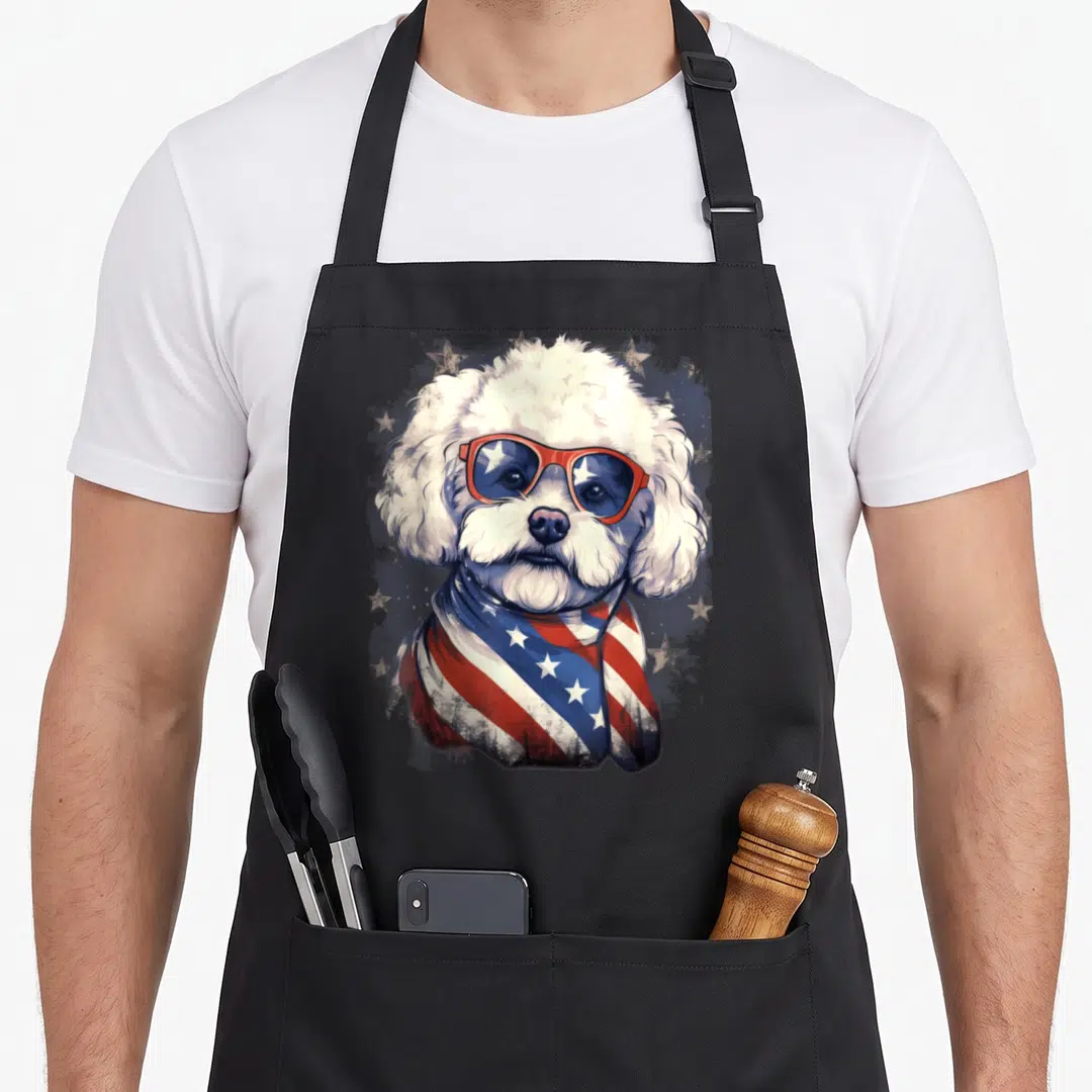 Bichon 4th of july apron - White dog with red sunglasses and US flag scarf on distressed background — shown on a black cotton canvas apron against a white background